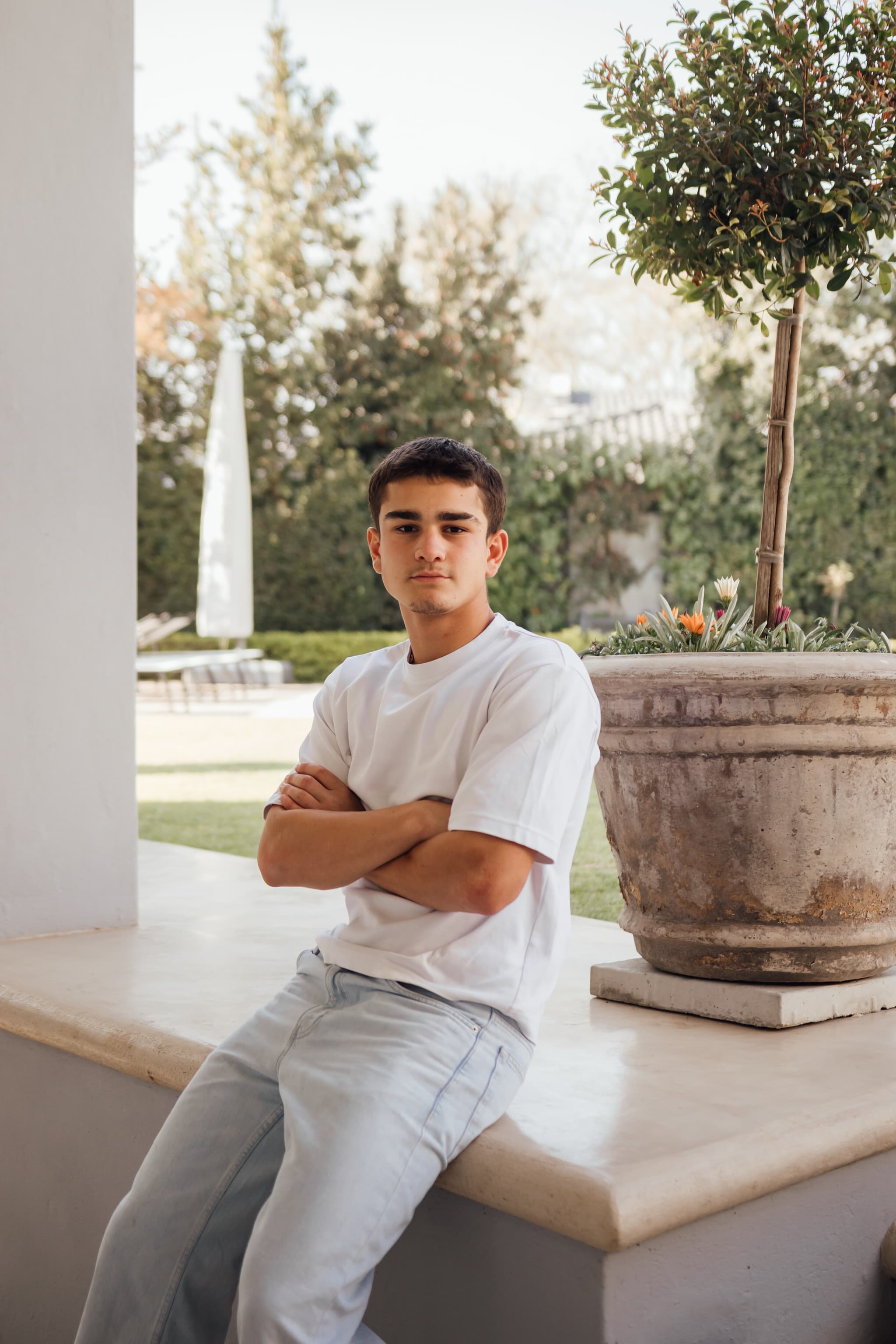 A model in a white tee and light wash jeans, arms crossed, sitting on a stone ledge beside a potted tree